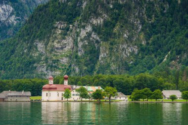 A white church with red domes stands by the tranquil waters of a lake, surrounded by lush greenery and majestic mountains on a sunny day. Konigssee lake Bavaria German Alps Berchtesgaden church by the clipart