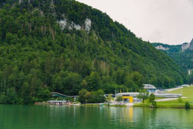 Vintage wooden boathouses line the calm, green waters, surrounded by dense trees and towering mountains under a cloudy sky, creating a tranquil atmosphere in nature. Konigssee lake Bavaria German Alps clipart