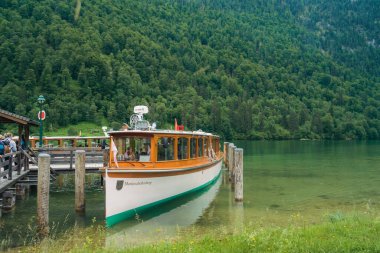 Bir tekne, bulutlu bir gökyüzünün altında, dramatik yeşil tepeler ve yoğun ormanlarla çevrili güzel bir gölün sakin sularında zarifçe hareket eder. Barışçıl ortam rahatlamaya davetiye çıkarıyor. Konigssee Bavyera