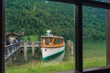 Bir tekne, bulutlu bir gökyüzünün altında, dramatik yeşil tepeler ve yoğun ormanlarla çevrili güzel bir gölün sakin sularında zarifçe hareket eder. Barışçıl ortam rahatlamaya davetiye çıkarıyor. Konigssee Bavyera