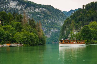 A boat gracefully moves through the calm waters of a beautiful lake, bordered by dramatic green hills and dense forests under a cloudy sky. The peaceful setting invites relaxation. Konigssee Bavaria clipart