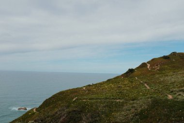 Cabo da Roca Sintra Portekiz 'deki manzaralı kıyı şeridi boyunca yürüyen yürüyüşçüler Atlantik Okyanusu' na bakan çimenli kayalıklarla Avrupa 'nın en batısındaki bulutlu gökyüzünün altında açık hava seyahatleri yapıyorlar. Yüksek