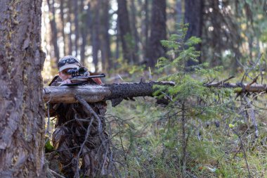 Kamuflaj kıyafetli avcı tüfeği doğrultup devrilmiş bir ağaca dayadı. Tüfek dürbününden bakıyorum. Çekime hazırız. Avlanma.
