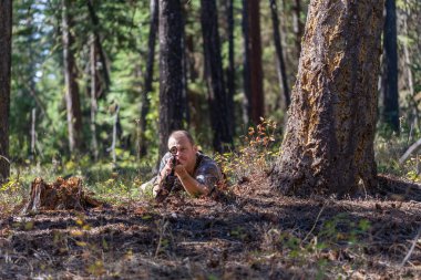 Kamuflaj elbiseli avcı ağacın altında yerde yatıyor, dürbünden bakarken tüfeği doğrultuyor. Çekime hazırız. Ön manzara. Avlanma.