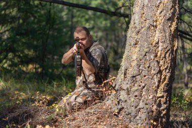 Kamuflaj elbiseli avcı ağaçların arkasında oturmuş dürbünden bakarken av tüfeğini doğrultuyor. Çekime hazırız. Ön görünüm