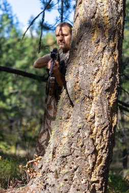 Avcı büyük bir ağacın arkasında saklanıyor ve önünde tüfeğini tutuyor. Çekime hazırız. Yüzündeki gölge