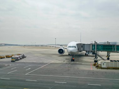 Pekin Başkenti Uluslararası Havalimanı 'na park edilmiş büyük bir yolcu uçağı, yanında bir jet köprüsü ve ön şaside çalışan bir servis ekibi var. Yüklü yük arabalarıyla bagaj römorkörü. Taksiler ve uzak terminal binaları.