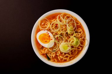 Baharatlı ramen eriştesinin üst görüntüsü, siyah bir arka planda sızıntı halkaları ve yarım yumurtayla portakal kırmızısı çorbasına batırılmış..