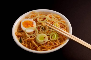 Kırmızı biberli çorba içinde yüzen erişteli ramen çorbası dairesel pırasa dilimleri ve yumurta ile süslenmiş, harekete hazır yemek çubukları. Siyah arkaplan.