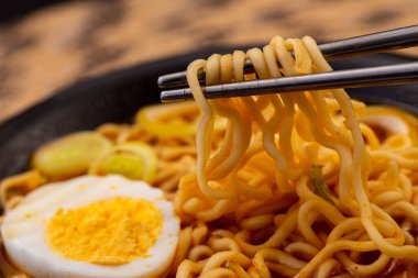 Dokuma hasırının üzerinde, dilimlenmiş pırasalarla süslenmiş, et suyuna bulanmış eriştenin yakın çekimi. Bir çift metal çubuk eriştenin bir kısmını kaldırıyor. Açı görünümü.