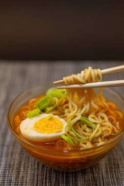 Hazır erişte porsiyonu, yemek çubuklarıyla, et suyuyla, dilimlenmiş pırasa ve yumurta ile süslenmiş cam bir kasede servis edilmiş gri paspasın üzerinde. Portre yönelimi.