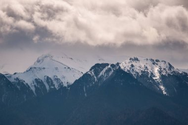 Romanya 'daki Bucegi Dağları' nın dramatik bulutlarla çevrelenmiş karla kaplı zirvelerinin büyüleyici bir görüntüsü. Engebeli dağ manzarası ve sakin kış manzarası burayı doğa severler, maceracılar ve fotoğrafçılar için mükemmel bir yer yapıyor.