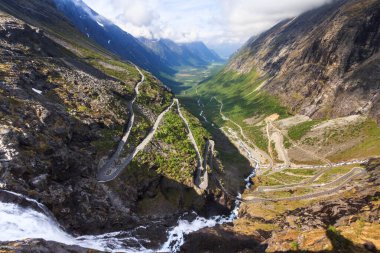 A breathtaking summer view of Trollstigen, Norway, with a cascading waterfall in the foreground and a winding valley stretching into the distance. clipart