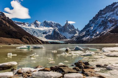 Göz kamaştırıcı Laguna Torre güneşin altında parıldıyor. Uzakta ikonik dağlar ve sakin sularında yüzen kırık buzlar.