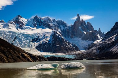 Güneşli bir günde Arjantin Patagonya 'daki Laguna Torre manzarası. Gölün berrak suları ikonik dağların güzelliğini yansıtırken, dağılmış buzdağları dramatik bir dokunuş ekliyor. Sahne, doğanın bu uzak diyardaki ihtişamının bir göstergesidir.