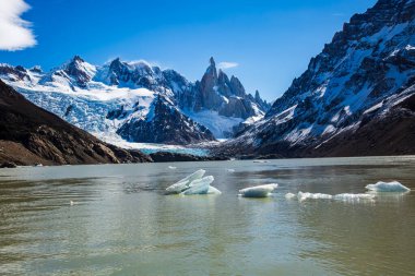 Göz kamaştırıcı Laguna Torre güneşin altında parıldıyor. Uzakta ikonik dağlar ve sakin sularında yüzen kırık buzlar.