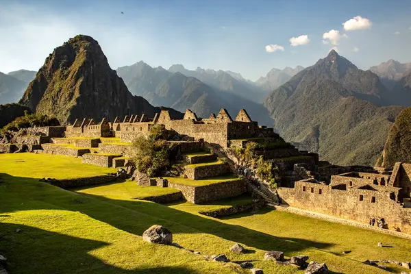 Bu fotoğraf, Peru 'daki Machu Picchu' nun dünyaca ünlü mekanını güneşli bir gökyüzünün altında gösteriyor. Eski kalıntılar, mükemmel şekilde korunan çimlerin yanında vurgulanırken, turistler merdivenlerden inerken minicik görünüyorlar. İkonik dağ geri döndü