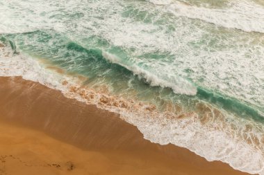 Avustralya 'da Büyük Okyanus Yolu' ndaki kumlu bir sahile düşen dalgaların hava görüntüsü. Ayak izleri kumu boydan boya geçiyor, manzaralı kıyı şeridine insan dokunuşu katıyor.