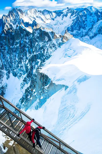 Ceketli iki turist, büyük karlı dağlarla çevrili Chamonix 'in yukarısındaki metal bir geçitte yürüyor. Güneş parlak bir gökyüzünün altında, tepelerde parlıyor..