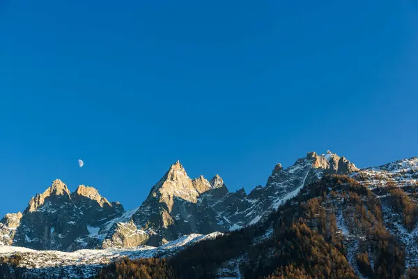 Güneşli bir kış gününde açık mavi gökyüzüyle Chamonix 'in engebeli tepelerinde ay batıyor. Karlı Alp manzarası.