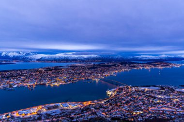 Kış mevsiminde Tromso, Norveç 'in alacakaranlık hava manzarası. Şehir ışıkları tamamen bulutlu gökyüzünün altında parıldıyor, fiyort üzerindeki köprü uzaklıktan görülebiliyor.