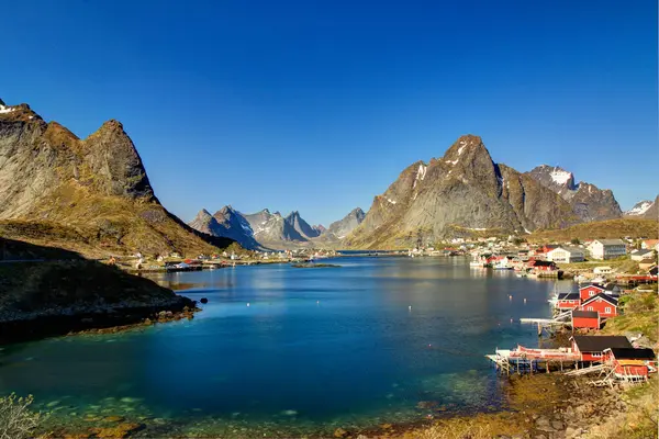 Reine, Lofoten, Norveç manzaralı, kırmızı balıkçı evleri ve yüksek dağları var. Fiyort suları güneşli bir gökyüzünün altında parıldıyor..