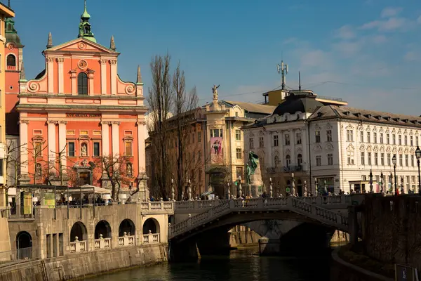Ljubljana 'nın Fransisken Kilisesi' nin (Ave, Gratia Plena) güneşli gündüz manzarası ikonik Üçlü Köprü 'nün (Tromostovje) yanında. Canlı bir kültürel dönüm noktası.