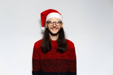 Uzun saçlı, Noel kırmızı kazaklı ve beyaz arka planda Noel Baba şapkalı genç adamın stüdyo portresi..