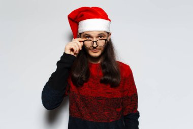 Uzun saçlı, gözlüklü, kırmızı Noel süveteri giyen ve beyaz arka planda Noel Baba şapkası takan genç bir adamın stüdyo portresi..