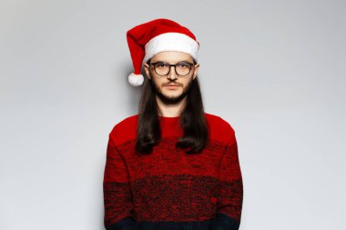 Uzun saçlı, gözlüklü, kırmızı Noel süveteri ve beyaz arka planda Noel Baba şapkası takan ciddi bir adamın stüdyo portresi..
