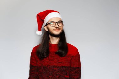 Uzun saçlı, gözlüklü, kırmızı Noel süveteri ve beyaz arka planda Noel Baba şapkası takan komik bir adamın stüdyo portresi..
