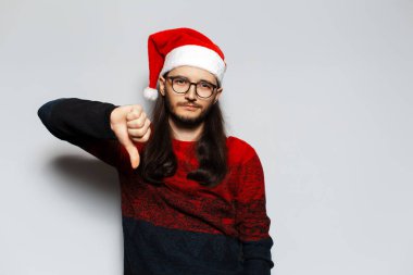 Uzun saçlı ve gözlüklü bir gencin stüdyo portresi. Kırmızı Noel süveteri ve beyaz arka planda Noel Baba şapkası takıyor..