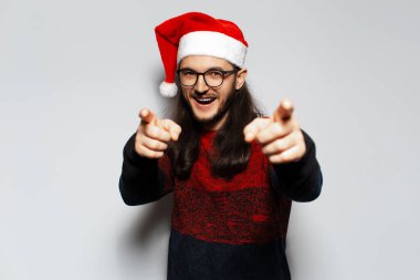 Uzun saçlı, gözlüklü, işaret parmağıyla seni gösteren mutlu genç adamın stüdyo portresi beyaz arka planda kırmızı Noel süveteri ve Noel Baba şapkası giyiyor..