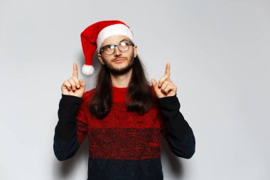Uzun saçlı, işaret parmağı havada, kırmızı Noel süveteri ve beyaz arka planda Noel Baba şapkası giyen yakışıklı bir adamın stüdyo portresi..