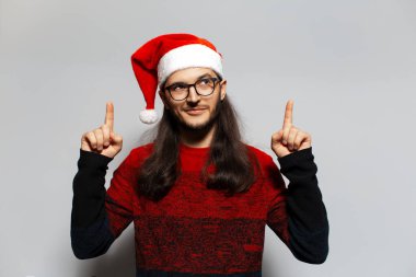 Uzun saçlı, işaret parmağı havada, kırmızı Noel süveteri ve beyaz arka planda Noel Baba şapkası giyen yakışıklı bir adamın stüdyo portresi..