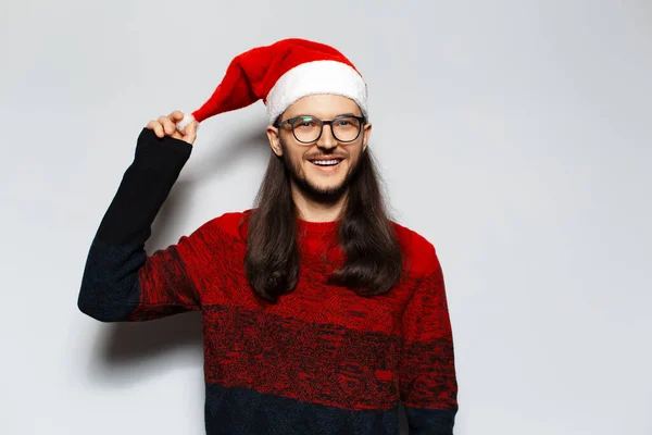 長い髪と眼鏡を持つ若い笑顔の男のスタジオの肖像画は 白い背景にサンタの帽子のポンポンを保持し 赤いクリスマスセーターを身に着けています — ストック写真