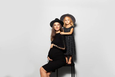 Stüdyoda karnına dokunan hamile bir kadının beyaz arka planda anne ile kızının portresi. Siyah şapkalı..