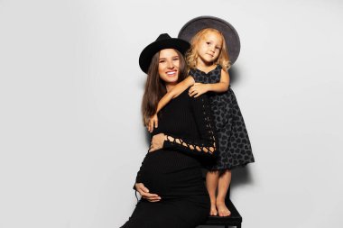 Genç hamile bir kadının stüdyo portresi, beyaz arka planda annesine sarılan bir kız. Siyah şapkalı..