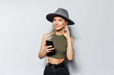 Kablosuz kulaklık takan, akıllı telefon kullanan, elinde cihazla beyaz arka planda gri şapka ve gömlek giyen sevimli, gülümseyen bir kızın stüdyo portresi..