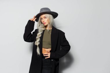 Saçları örgülü, siyah ceketli ve gri şapkalı, beyaz arka planda güzel sarışın bir kızın stüdyo portresi..