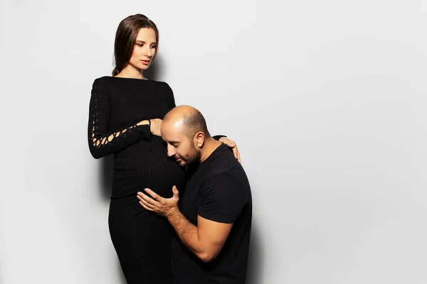 Studio portrait of young couple, future parents. Pregnant woman, young father touching the belly of his wife on white background. Wearing black.