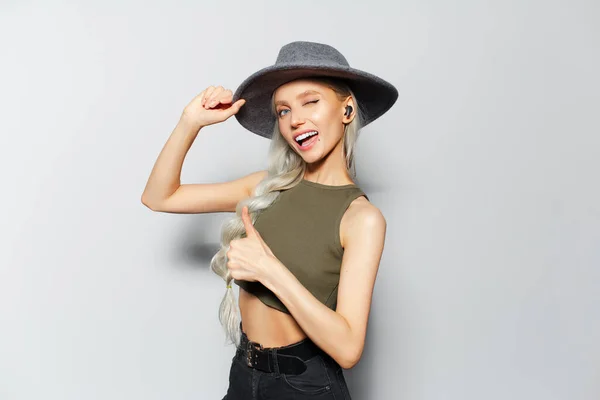 Studio portrait of happiness blonde girl with wireless earphones in ears, showing thumbs up, on white background. Wearing grey hat, green shirt and black jeans.