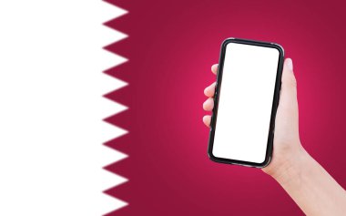 Close-up of male hand holding smartphone with blank on screen, on background of blurred flag of Qatar.