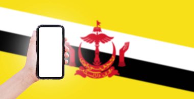 Ekranda boş, Brunei 'nin bulanık bayrağının arka planında akıllı telefonu tutan bir erkek. Yakın çekim görünümü.