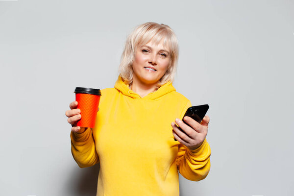 Studio portrait of blonde woman using smartphone, holding paper cup for coffee takeaway of red color, wearing yellow sweater on white background.