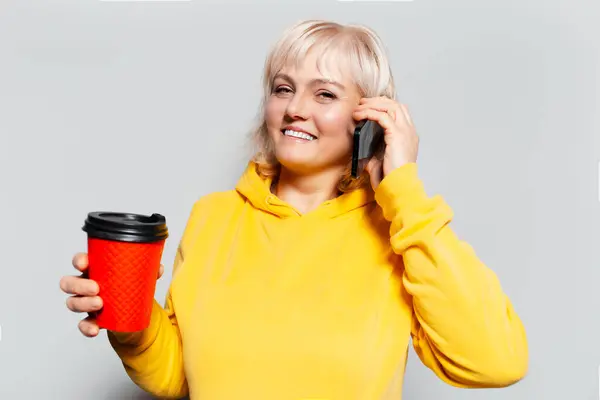 Студийный Портрет Улыбающейся Блондинки Говорящей Смартфоне Держащей Бумажную Чашку Кофе — стоковое фото