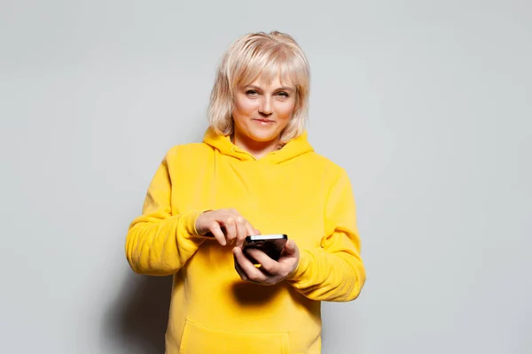 穿着黄色毛衣的金发女人用白色背景智能手机的工作室肖像 — 图库照片