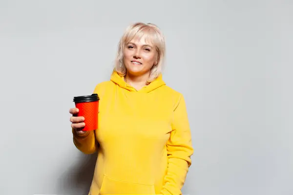 身着黄色毛衣 拿着红纸杯在白色背景下外卖咖啡的金发微笑女子的工作室肖像 — 图库照片