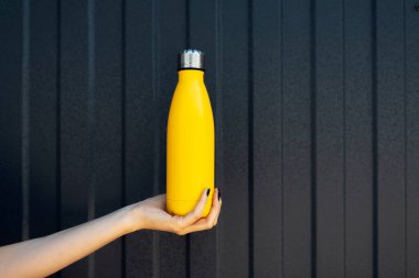 Metal zemin üzerinde siyah çizgileri olan sarı renkli bir termosu olan kadın elinin yakın plan fotoğrafı..