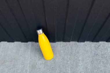 Çelik termik su şişesinin üst görüntüsü. Beton zemin üzerinde sarı renkli. Metal arka plan ise siyah renkte..